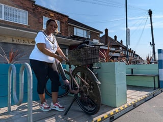 Smiling woman with her bicycle, hand on the handlebars and a wicker basket on the front, standing on a raised wooden platform beside planters and benches outside neighbourhood shops.