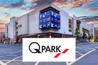 The Q-Park logo sitting in front of one of their multi storey car parks with signage on the outside relating to the facilities inside